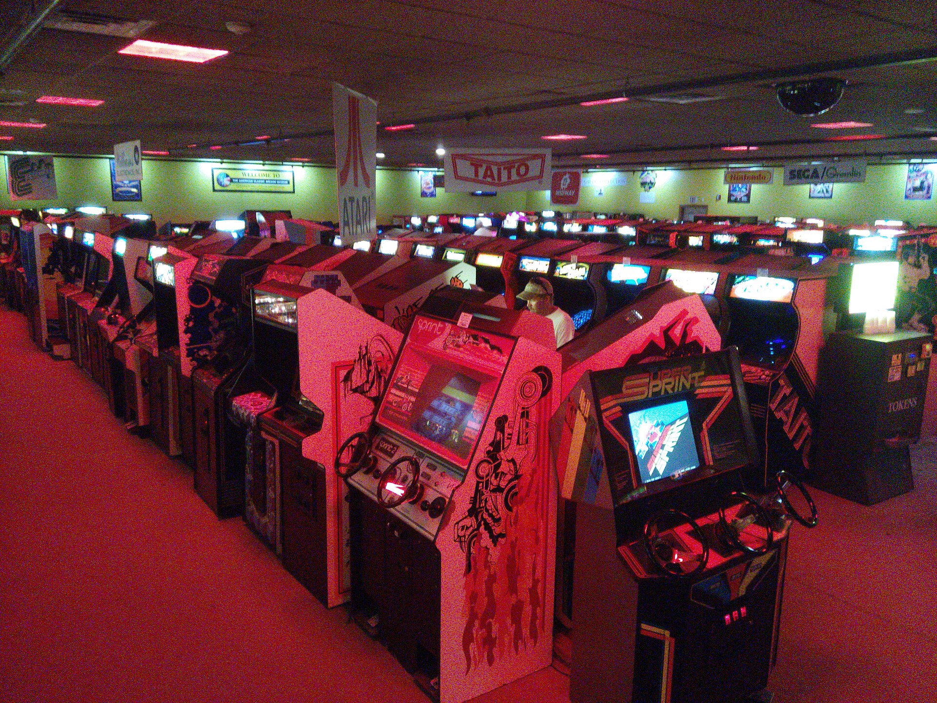Rows of 1980s arcade cabinets under red light, with Atari and Taito banners hanging above