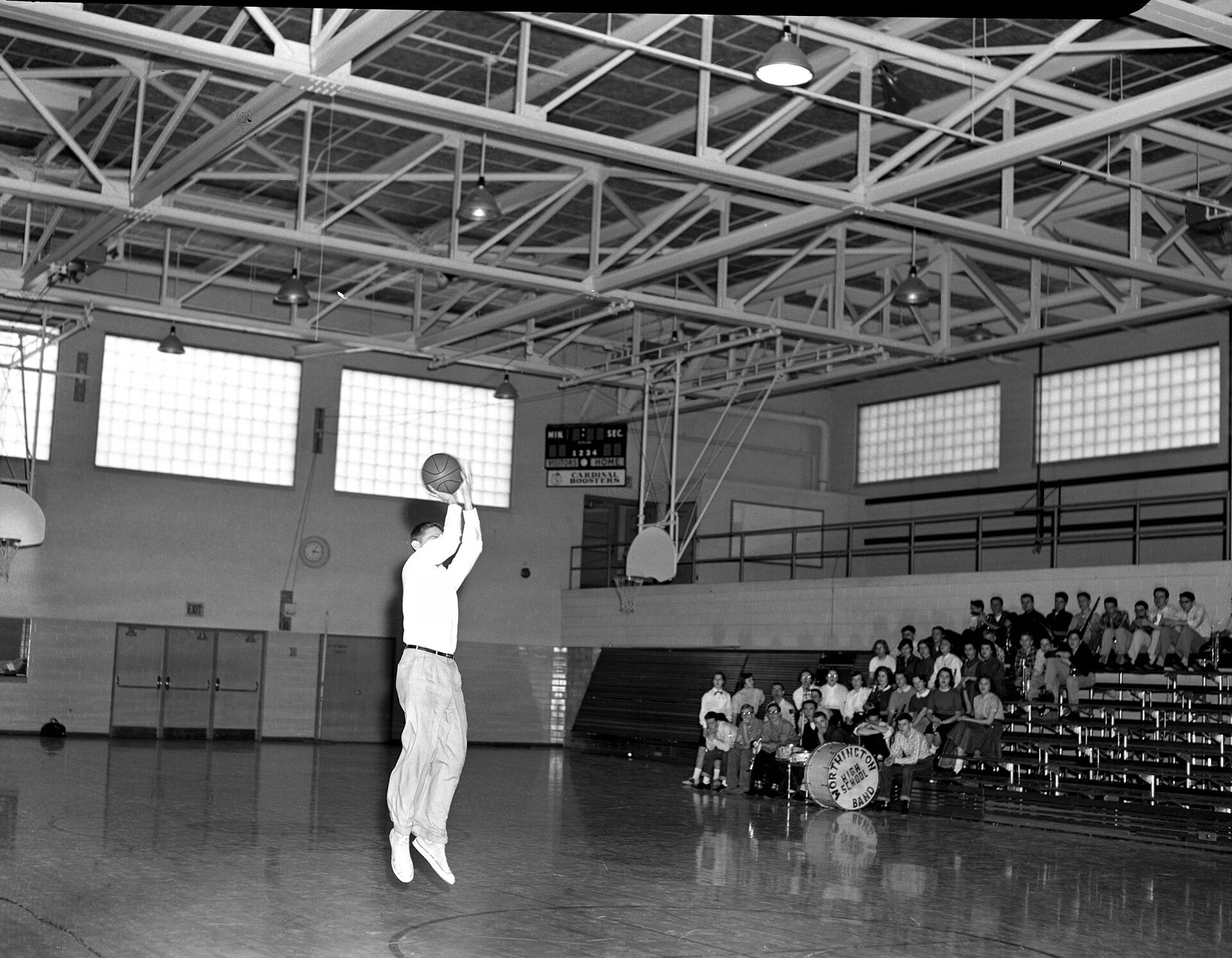 A high school player in a white shirt leaps with the ball above his head in an empty 1950s gym while the band watches from the stands